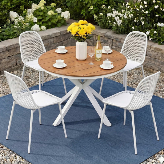 Outdoor dining set with a round wooden table and four white chairs on a blue rug, surrounded by flowers and a stone wall.