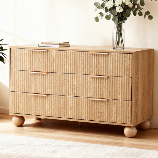 Wooden dresser with three drawers on a wooden floor, featuring a vase of flowers on top.